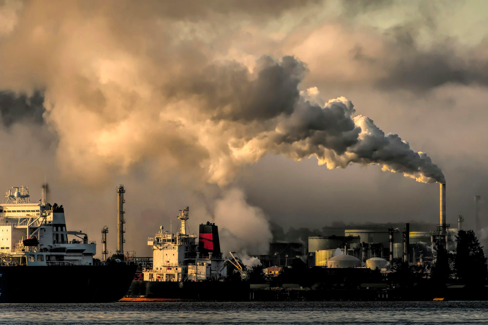 A refinery and tanker terminal silhouetted against a smoke-lit sky at dusk